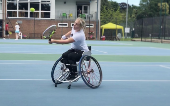 Ruby is on a blue outdoor tennis court. She is playing wheelchair tennis, hitting the ball with her racket.