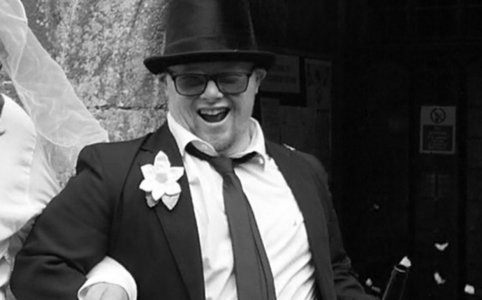 A black and white image of a man in a suit and top hat wearing glasses, with his arm around a bride. 